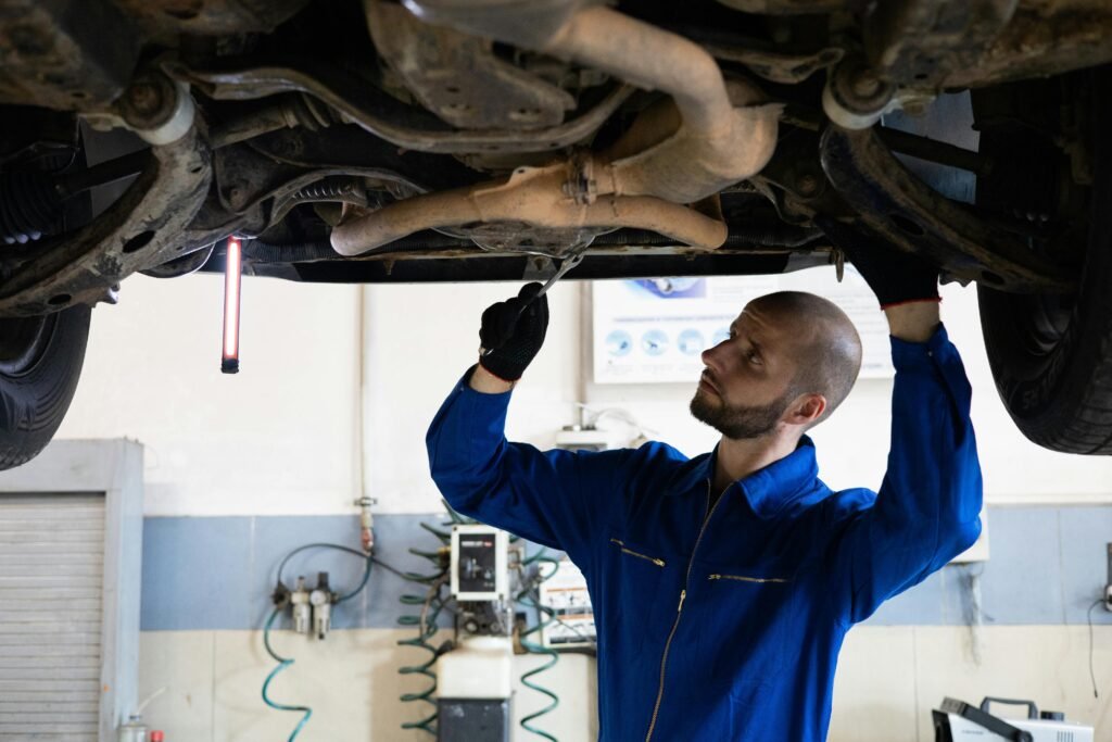 pexels-photo-8986100-8986100 A mechanic in blue overalls inspects the exhaust system of a car in a workshop.